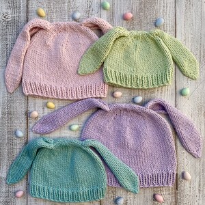 May include: Four knitted bunny hats in pink, green, lavender, and a lighter shade of green. The hats have long bunny ears and are arranged on a white wooden surface with pastel colored Easter eggs scattered around them.