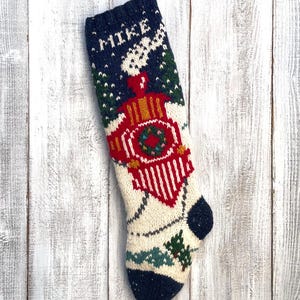 May include: A knitted Christmas stocking featuring a train design. The stocking has a cream-colored body with a navy blue cuff and toe. The train is red and gold, with a green wreath. The name "MIKE" is knitted at the top.
