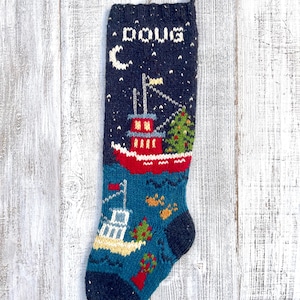 Può includere: Una calza natalizia lavorata a maglia blu scuro con il nome DOUG in lettere bianche nella parte superiore. Il design presenta una barca rossa con un albero di Natale verde, una luna crescente bianca e una barca bianca più piccola su un mare verde acqua.