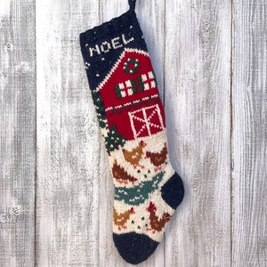 Peut inclure: Une chaussette de Noël en tricot avec une grange rouge, des arbres verts et des poulets blancs sur un fond blanc. La chaussette est bleue et blanche avec le mot "NOEL" tricoté dans le tissu.