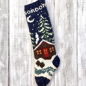May include: A navy blue knitted Christmas stocking with a winter cabin scene. The stocking features a cabin, trees, and a crescent moon against a starry sky. The name "GORDON" is knitted at the top. The stocking is approximately 51 cm long.