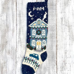 Peut inclure: Une chaussette de Noël tricotée bleu marine représentant une maison blanche avec une couronne sur la porte, un croissant de lune et le texte FAM en haut. Le motif inclut une barrière blanche, une voiture bleue et une cheminée avec de la fumée.