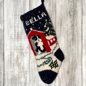 Könnte beinhalten: Ein blauer, gestrickter Weihnachtsstrumpf mit einer Winterszene und dem Namen "BELLA" obenauf gestickt. Der Strumpf zeigt eine rote Scheune, einen weißen Hund und eine verschneite Landschaft.