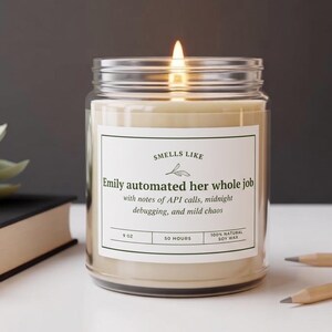 May include: A clear glass jar candle with a lit wick and a white label that reads: SMELLS LIKE Emily automated her whole job with notes of API calls, midnight debugging, and mild chaos. The label also states 9 oz, 50 hours, and 100% natural soy wax.