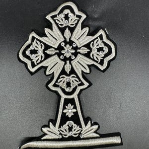 Puede incluir: Un parche de tela negra con forma de cruz ornamentada, que presenta un intrincado bordado metálico plateado y pequeñas lentejuelas transparentes en todo el diseño. La cruz tiene una base ensanchada y una barra horizontal en la parte inferior.