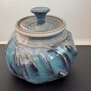May include: A ceramic jar with a lid featuring a swirling glaze of deep blue, turquoise, and white. The jar has a rounded, textured body with vertical indentations and a matching lid with a central knob handle, showcasing a glossy finish.