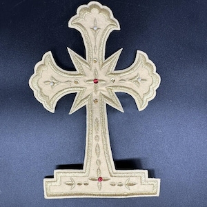 Può includere: Un'applicazione a forma di croce in tessuto beige con intricati ricami dorati in un motivo floreale e a stella. Il design è arricchito da piccoli strass rossi, dorati e bianchi incastonati su tutta la superficie, creando un emblema religioso decorativo.