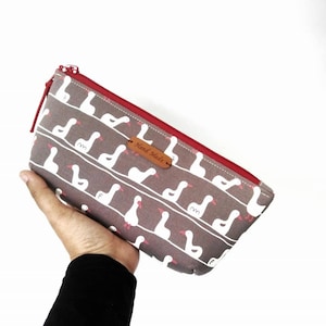 May include: A grey zipped pouch with a red zipper and a white goose pattern. The pouch has a brown leather tag that says "Hand Made".