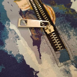 May include: A close-up of a zipper on a blue and white patterned fabric. The zipper is silver and has a gold pull tab.