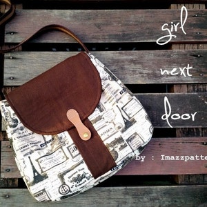 May include: A brown and white fabric crossbody bag with a flap closure and a brown leather strap. The bag has a vintage Parisian theme with a print of old French advertisements and illustrations. The bag is on a wooden surface with the text "girl next door by: Imazpatterns" written on it.