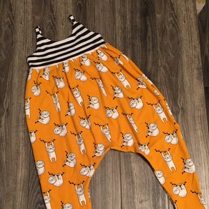 May include: A drop-crotch jumpsuit featuring a vibrant orange base with a repeating pattern of white sloths hanging from black branches. The garment includes a black and white horizontal striped bodice with thin straps and matching striped ankle cuffs.