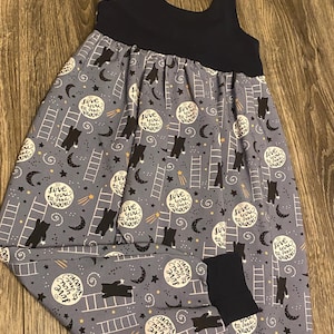 May include: A sleeveless jumpsuit with a dark navy bodice and a light blue patterned lower section. The pattern features black bears, white ladders, crescent moons, stars, and circular text that reads "love you to the moon".