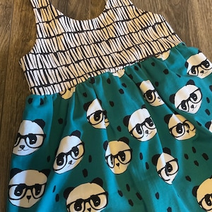 May include: A sleeveless dress featuring a bodice with a black and white vertical dash pattern and a gathered skirt in teal green. The skirt is decorated with a repeating print of white panda faces wearing black glasses and small black dots.
