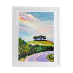 May include: A framed landscape painting shows a winding road leading up a green hill with trees. The sky has pastel pink, blue, and yellow hues. The artwork is enclosed in a white frame.