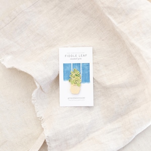 May include: A gold enamel pin featuring a potted fiddle leaf plant. The pin is on a white card with the text "The Zen Succulent Fiddle Leaf enamel pin @thezensucculent thezensucculent.com"