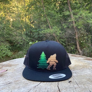 Puede incluir: Una gorra de camionero negra con un diseño bordado de un pino y un Bigfoot en verde y marrón. La gorra tiene una parte trasera de malla negra y un cierre de presión negro.