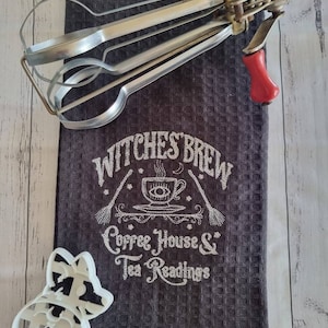 May include: A dark gray waffle-weave kitchen towel with the embroidered text "Witches Brew Coffee House & Tea Readings." An antique egg beater and a pig-shaped cookie cutter are also present.