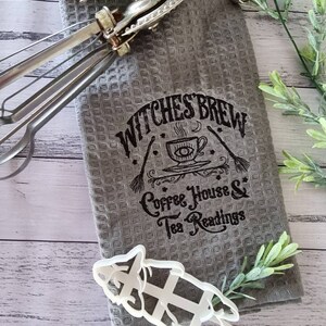 May include: A gray waffle-weave kitchen towel with the text "Witches Brew Coffee House & Tea Readings" in black. A vintage egg beater with red handles and a white cookie cutter are also present.