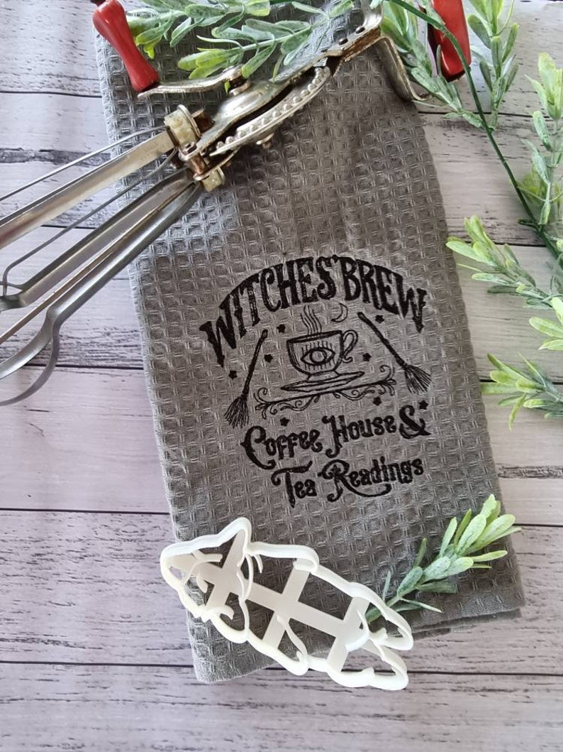 May include: A gray waffle-weave kitchen towel with the text "Witches Brew Coffee House & Tea Readings" in black. A vintage egg beater with red handles and a white cookie cutter are also present.