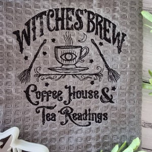 May include: Gray waffle-weave kitchen towel with black text and illustrations. The text reads "Witches Brew Coffee House & Tea Readings." A cup with an eye design, brooms, and stars are also featured.