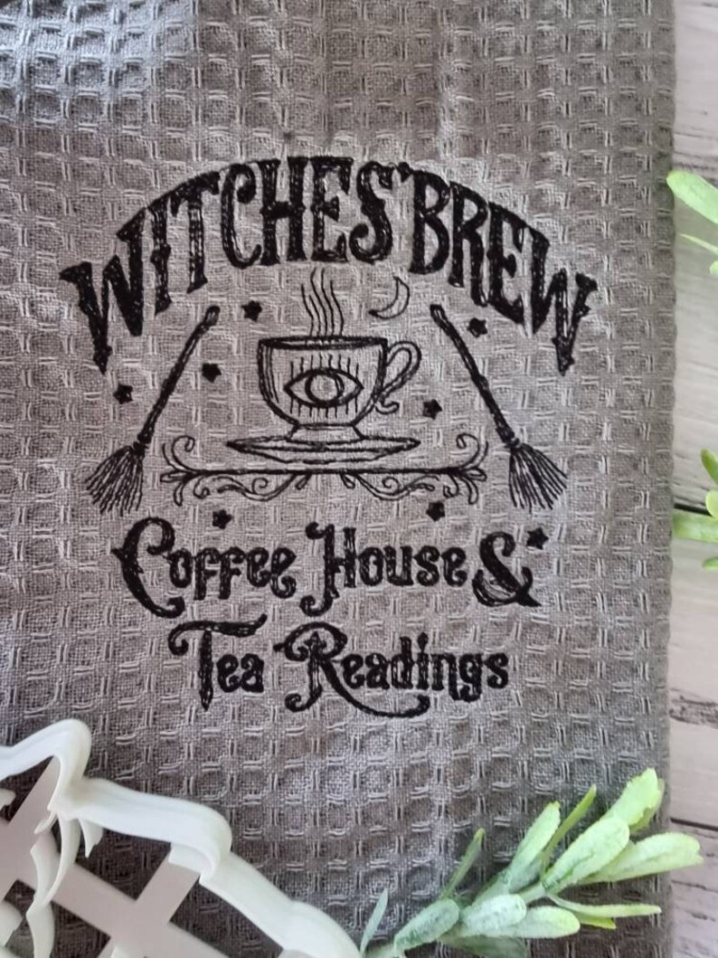 May include: Gray waffle-weave kitchen towel with black text and illustrations. The text reads "Witches Brew Coffee House & Tea Readings." A cup with an eye design, brooms, and stars are also featured.