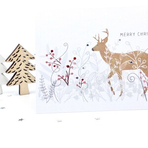 Op de afbeelding: Een witte kerstkaart met een gouden hert-illustratie en de tekst "Merry Christmas" in zwart. Het hert is omringd door zilveren en rode bessen en bladeren.