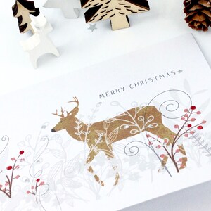 Op de afbeelding: Een witte kerstkaart met een gouden hert in een boslandschap. De kaart heeft de tekst "Prettige Kerst".