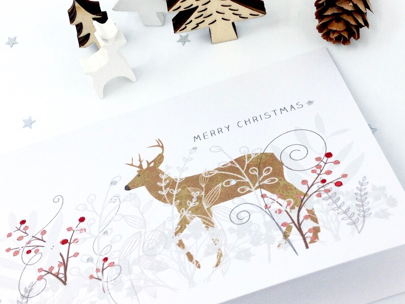 Op de afbeelding: Een witte kerstkaart met een gouden hert in een boslandschap. De kaart heeft de tekst "Prettige Kerst".