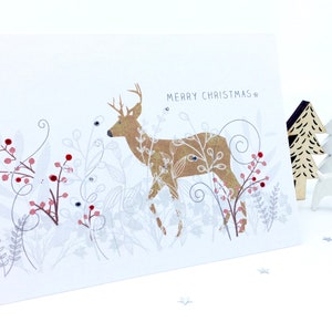 Op de afbeelding: Een gouden hinde met witte ogen en geweien staat in een wit en zilveren bloemenontwerp met rode bessen. De tekst "Merry Christmas" staat in het zwart bovenaan de kaart.