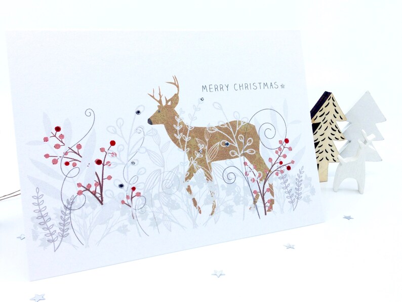Op de afbeelding: Een gouden hinde met witte ogen en geweien staat in een wit en zilveren bloemenontwerp met rode bessen. De tekst "Merry Christmas" staat in het zwart bovenaan de kaart.