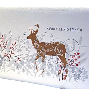 Op de afbeelding: Een witte kerstkaart met een bruin hert dat in een veld met zilver en rood blad staat. De tekst "Merry Christmas" staat in blauw bovenaan de kaart.