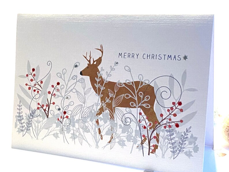 Op de afbeelding: Een witte kerstkaart met een bruin hert dat in een veld met zilver en rood blad staat. De tekst "Merry Christmas" staat in blauw bovenaan de kaart.