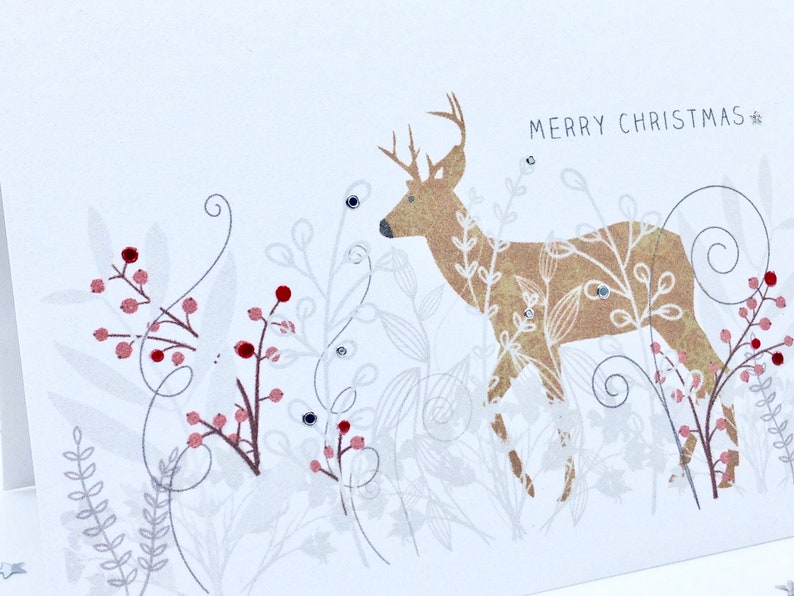 Op de afbeelding: Een witte kerstkaart met een gouden hinde-illustratie en de tekst "Merry Christmas". De hinde is omringd door rode bessen en wit blad.