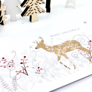 Op de afbeelding: Een witte kerstkaart met een gouden hert-illustratie omringd door zilveren en rode bessen en bladeren. De tekst "Merry Christmas" is in zwart bovenaan de kaart gedrukt.