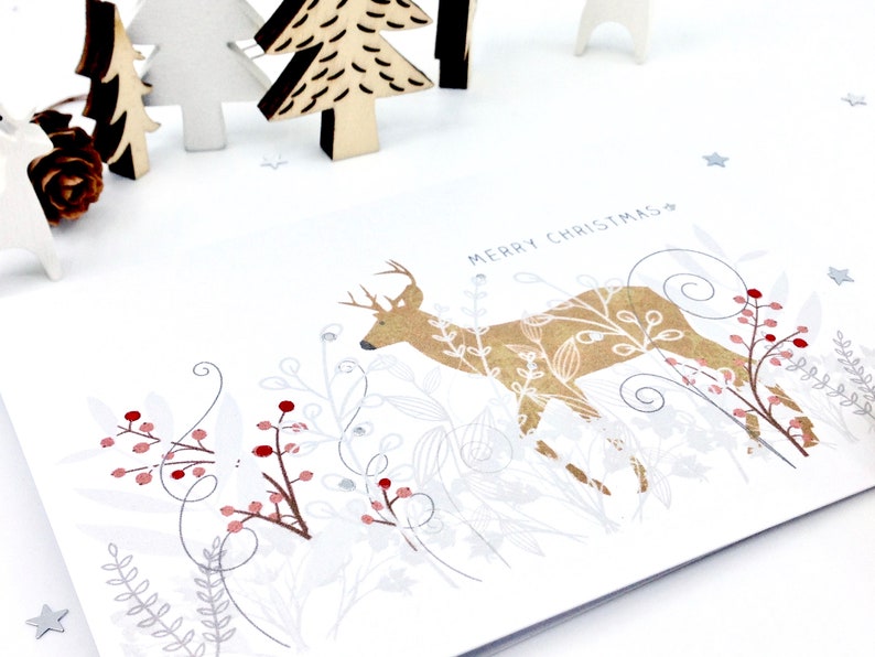 Op de afbeelding: Een witte kerstkaart met een gouden hert-illustratie omringd door zilveren en rode bessen en bladeren. De tekst "Merry Christmas" is in zwart bovenaan de kaart gedrukt.