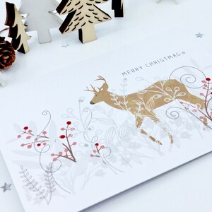 Op de afbeelding: Een witte kerstkaart met een gouden hert dat door een bos met zilveren en rode bessen loopt. De tekst "Merry Christmas" staat op de kaart gedrukt.