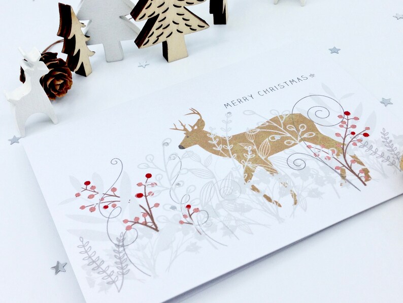 Op de afbeelding: Een witte kerstkaart met een gouden hert dat door een bos met zilveren en rode bessen loopt. De tekst "Merry Christmas" staat op de kaart gedrukt.