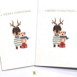 Puede incluir: Dos tarjetas de Navidad con una ilustración de perro. Uno lleva cuernos y un suéter a rayas, el otro una bufanda azul. Ambos sostienen un regalo envuelto. Las palabras "MERRY CHRISTMAS" están impresas en la parte superior.