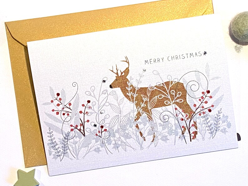 Op de afbeelding: Een witte kerstkaart met een gouden hert dat in een bos van zilveren en rode bessen staat. De kaart heeft de tekst "Merry Christmas".