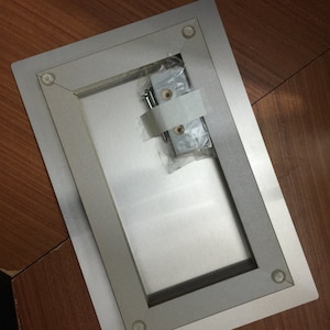 May include: A silver metal frame with a rectangular opening. The frame has four small, clear plastic bumpers on the corners. A small plastic bag containing two screws is visible inside the frame.