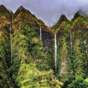 May include: A lush green mountainside with multiple waterfalls cascading down the rock face. The waterfalls are surrounded by dense foliage and mist.