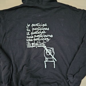 May include: Black hoodie with white text that says "Je participe tu participes il participe nous participons vous participerez ils profitent" and a drawing of a hand holding a paintbrush.