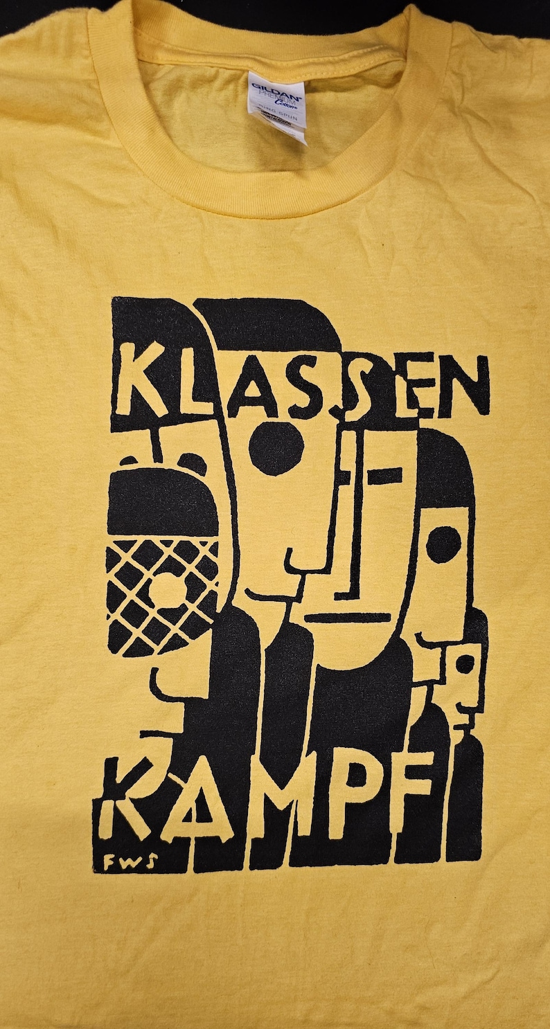 May include: Yellow t-shirt with a black graphic design of abstract faces and the text "KLASSEN KAMPF FWS".