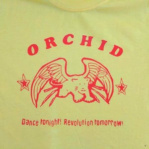 May include: A bright yellow t-shirt featuring a red graphic print. The design includes the word ORCHID in an arc above a winged feline creature flanked by two stars. Below the graphic, the text reads: Dance tonight! Revolution tomorrow!