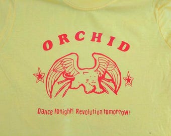 May include: A bright yellow t-shirt featuring a red graphic print. The design includes the word ORCHID in an arc above a winged feline creature flanked by two stars. Below the graphic, the text reads: Dance tonight! Revolution tomorrow!