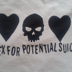 Könnte beinhalten: Weißes T-Shirt mit einem schwarzen Totenkopf und zwei schwarzen Herzen, die auf der Vorderseite aufgedruckt sind. Der Text "Index for Potential Suicide" ist unter dem Motiv aufgedruckt.