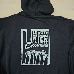 May include: Black hooded sweatshirt with a white graphic print on the back. The graphic features the text "LE VOTE NE CHANGE RIEN LA LUTTE CONTINUE" and a series of raised fists.