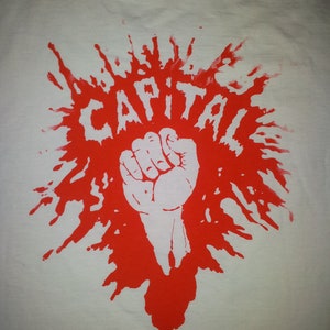May include: A white t-shirt with a red splatter design. The design features a raised fist and the word "CAPITAL" in red.