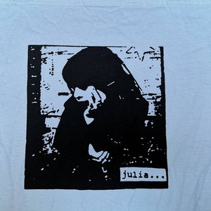 Può includere: Immagine in bianco e nero di una persona con la testa bassa, stampata su una maglietta bianca. Il testo "julia..." è stampato sotto l'immagine.