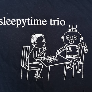 May include: A black and white illustration of a robot and a person playing a game on a table. The text "sleepytime trio" is at the top of the image.